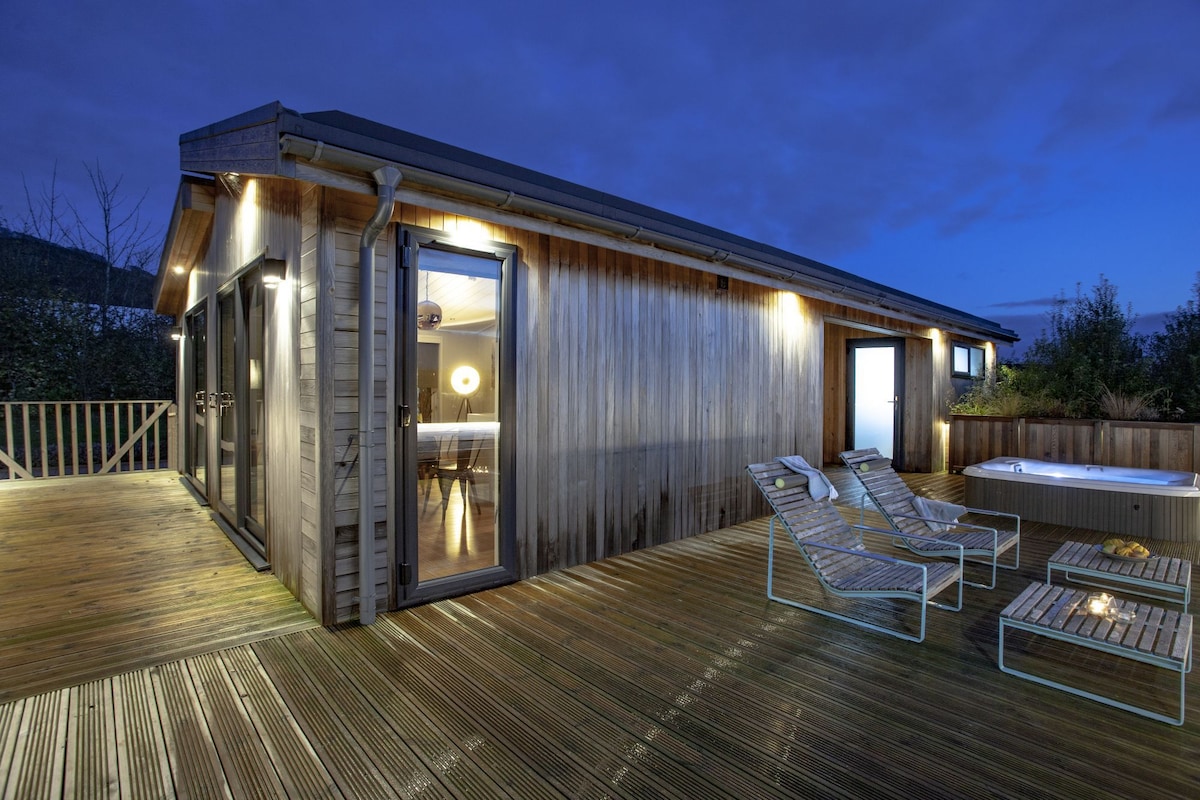 The exterior of the lodge is shown during dusk, featuring a spacious deck with led lighting. Two lounge chairs and a coffee table are visible, along with a partially sunken hot tub. Large glass doors offer views into the interior space.