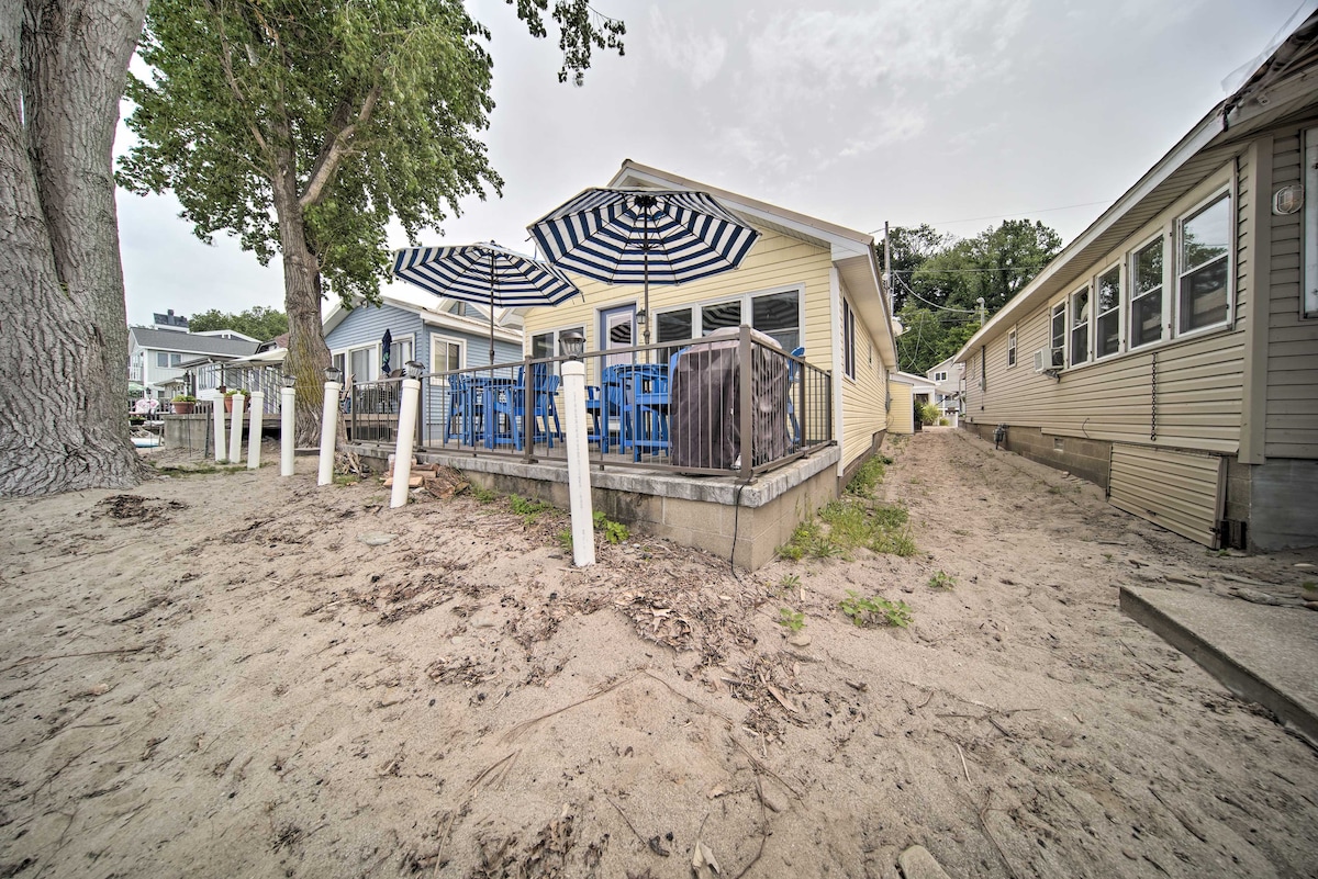 A sandy pathway leads to the cottage's outdoor patio area, which features blue chairs and striped umbrellas providing shade. The structure exhibits a casual beachside design, with nearby trees creating a natural border between properties.