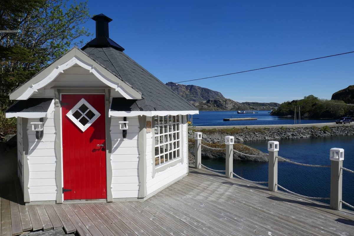 A charming wooden hut with a bright red door and diamond-shaped window accents is situated by the water. The surrounding wooden deck features lanterns, while scenic views of the shoreline and hills are visible in the distance, under a clear blue sky.