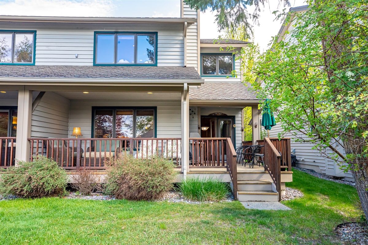 The exterior of the condo features a welcoming front porch with seating, surrounded by green grass and shrubs. Large windows allow natural light to fill the space. A patio table and umbrella provide an inviting outdoor area for relaxation.