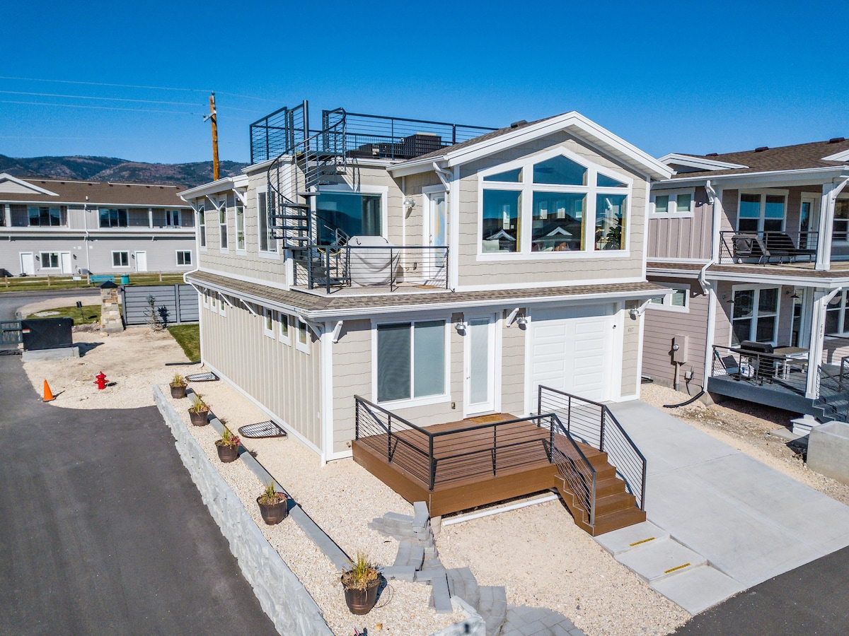 A modern two-story villa features a welcoming entrance with a wooden deck leading up to the door. Large windows illuminate the exterior, while a rooftop lounge area offers expansive views. The landscaped yard includes stone accents and flower pots along the pathway.