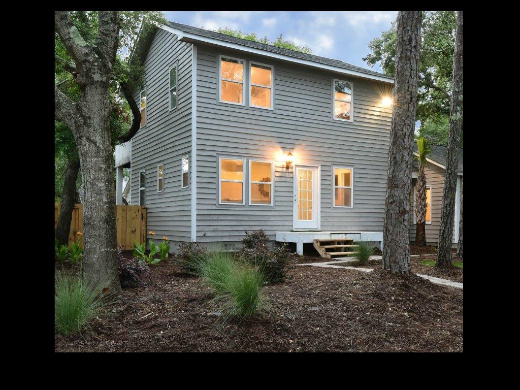 A two-story gray duplex features large windows that allow natural light to fill the exterior. A welcoming front porch leads to the main entrance, surrounded by landscaped greenery and mulch paths. Tall trees provide shade, enhancing the tranquil ambiance of the surroundings.