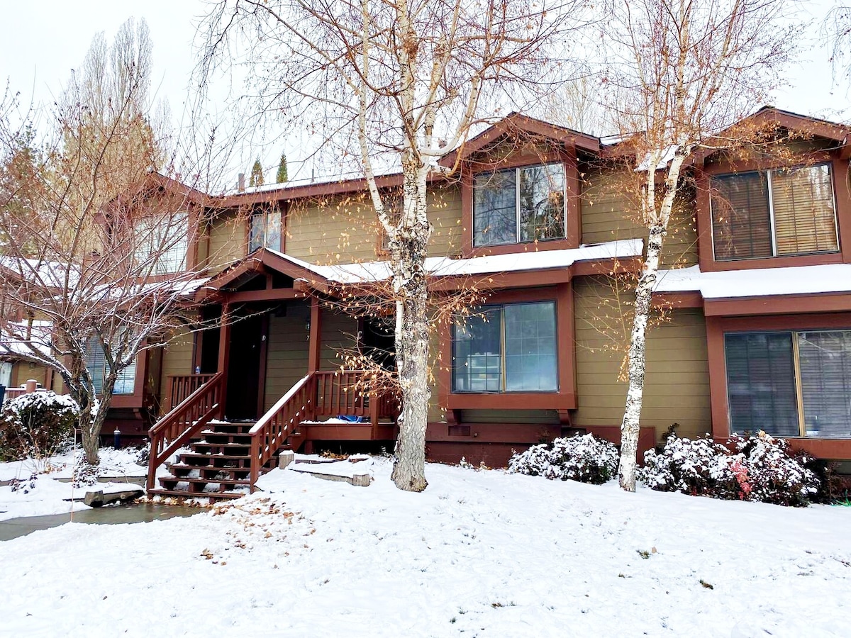A welcoming exterior view of a two-story retreat surrounded by a snow-covered landscape. The building features a wooden entry staircase leading to a porch. Leafless trees frame the property, and soft lighting accentuates the structure's design.
