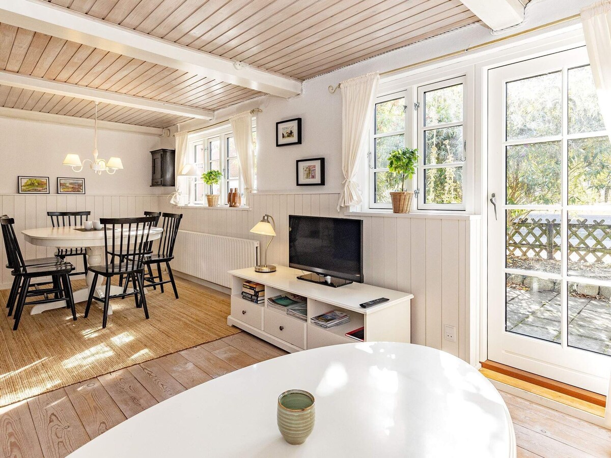 An inviting living and dining area features a light color palette with wooden beams and a woven rug. A round dining table is surrounded by black chairs, while a spacious TV unit stands across from a comfortable seating arrangement. Large glass doors open to a sunlit courtyard.