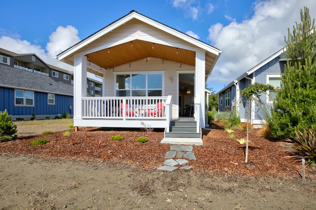 The cottage exterior is presented, featuring a welcoming front porch with white railings and comfortable seating. Neatly landscaped grounds surround the cottage, highlighted by a mulch bed and small plants. Clear skies with scattered clouds provide a bright, cheerful backdrop.