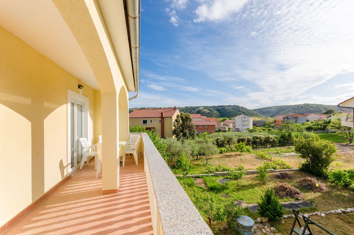 A spacious balcony is depicted, offering a view of lush greenery and distant hills. Sunlight brightens the area, highlighting a few white outdoor chairs, creating a calm setting for relaxation.