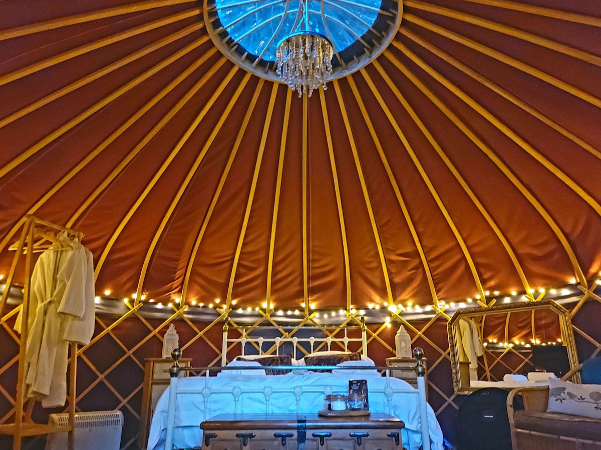 The interior of the yurt features a spacious bed adorned with soft linens, framed by warm fairy lights. A unique chandelier hangs from the center, illuminating the vintage decor. Comfortable robes are provided, creating a cozy retreat beneath the canvas ceiling.