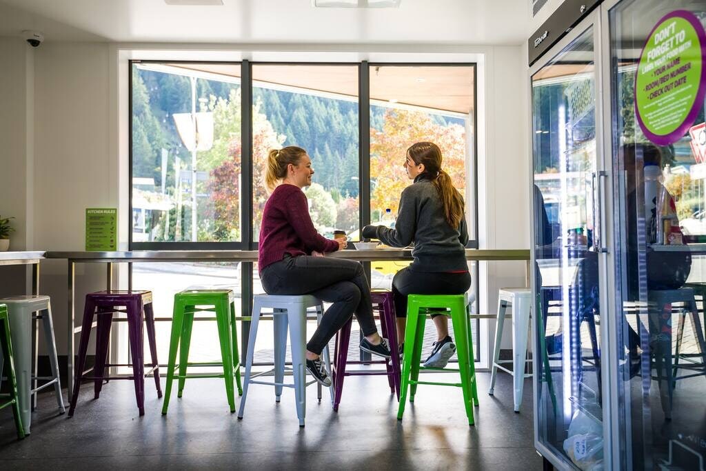 A bright communal space features two individuals seated at a high table. Colorful stools in purple and green add vibrancy to the room, which is enhanced by large windows allowing natural light to illuminate the area. Scenic views of trees and mountains are visible outside.