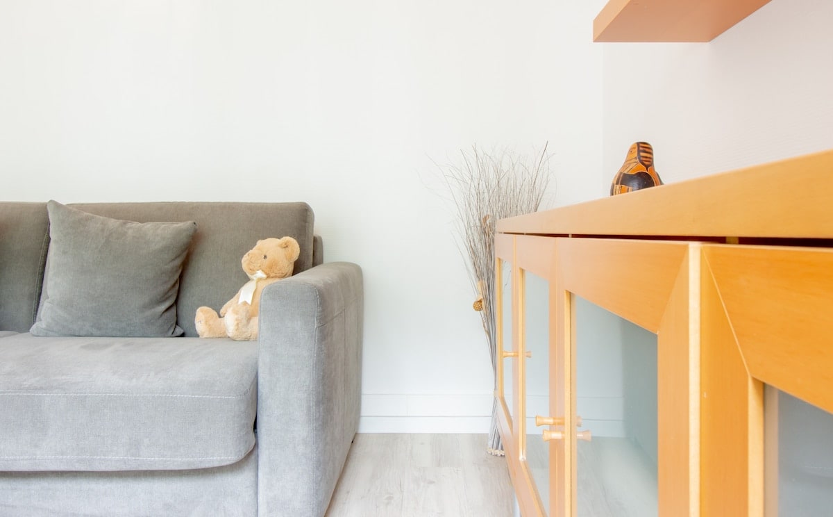 A comfortable gray sofa is positioned alongside a modern wooden sideboard. A plush teddy bear rests on the sofa, contributing to a cozy ambiance. Simple decorative elements are placed across the sideboard, enhancing the room's clean and airy design.
