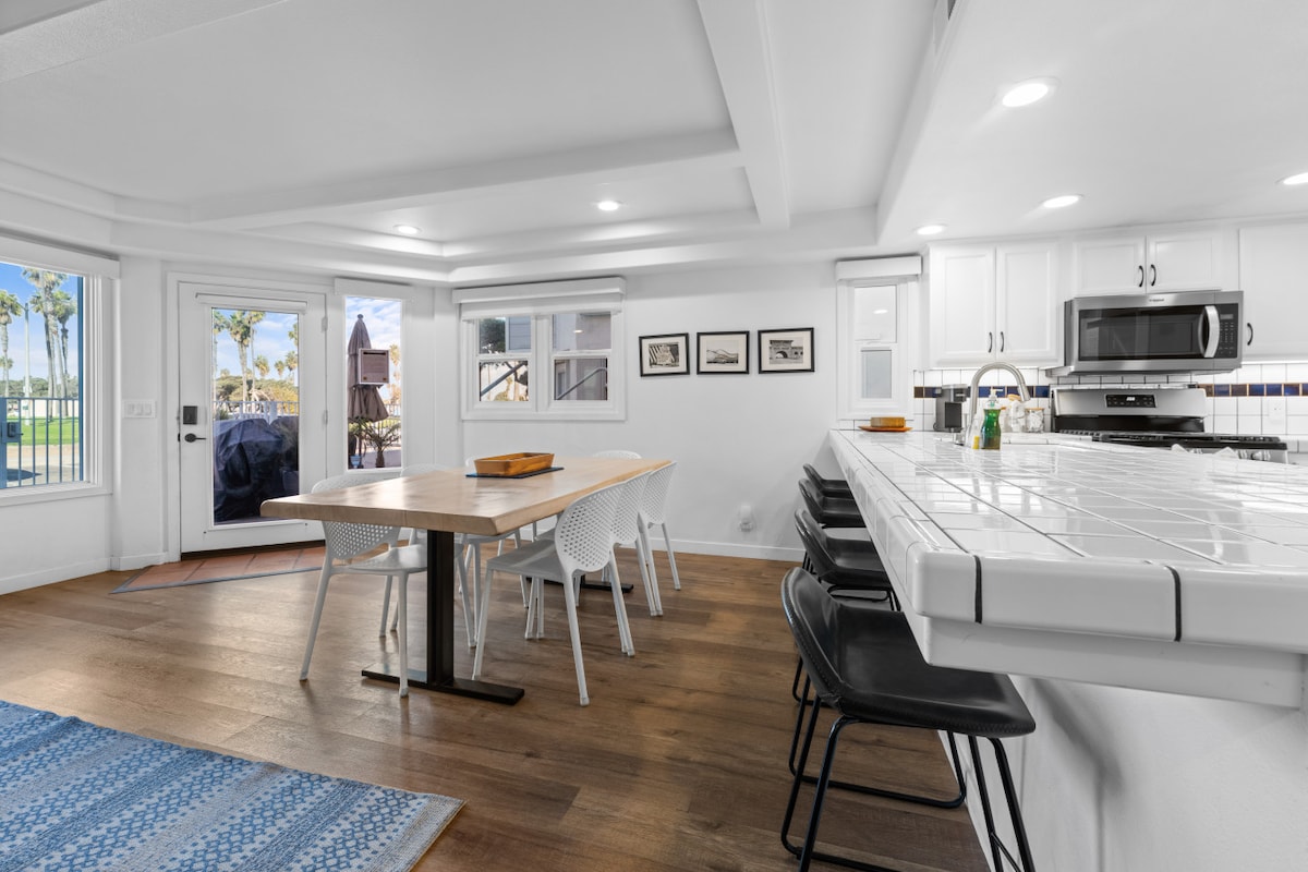 A bright dining area is complemented by a wooden table surrounded by white chairs, set against a backdrop of large windows that invite natural light. A modern kitchen with stainless steel appliances is visible, featuring a tiled countertop and minimalistic decor.