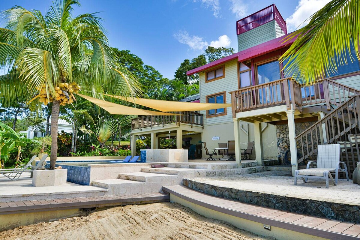 The exterior of a spacious rental home is showcased, featuring multiple levels with wooden decks. A vibrant canopy provides shade over a seating area, while palm trees and lush greenery surround the property. A pool area and sandy path lead toward the beach in the background.