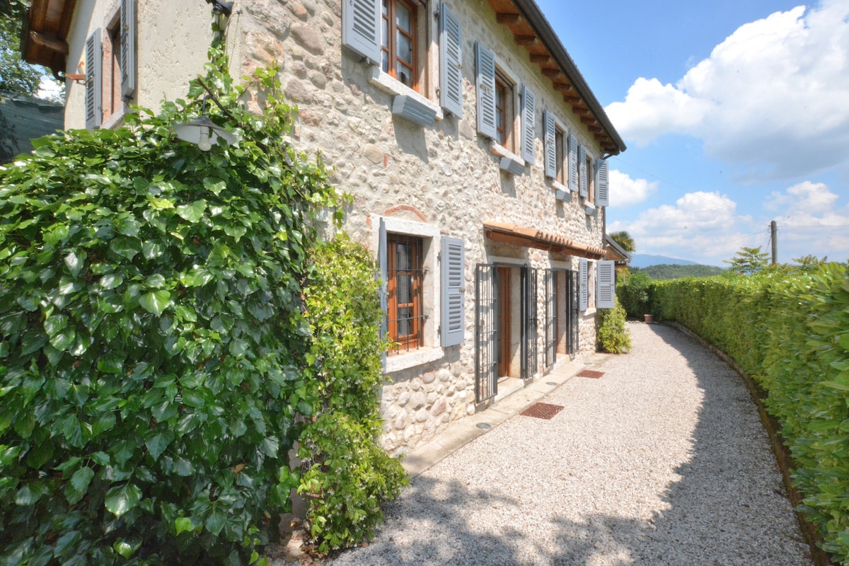 The exterior of the villa features a charming stone façade, adorned with wooden shutters and framed by lush greenery. A gravel pathway leads to the entrance, surrounded by tall hedges, creating a sense of privacy in this tranquil setting.