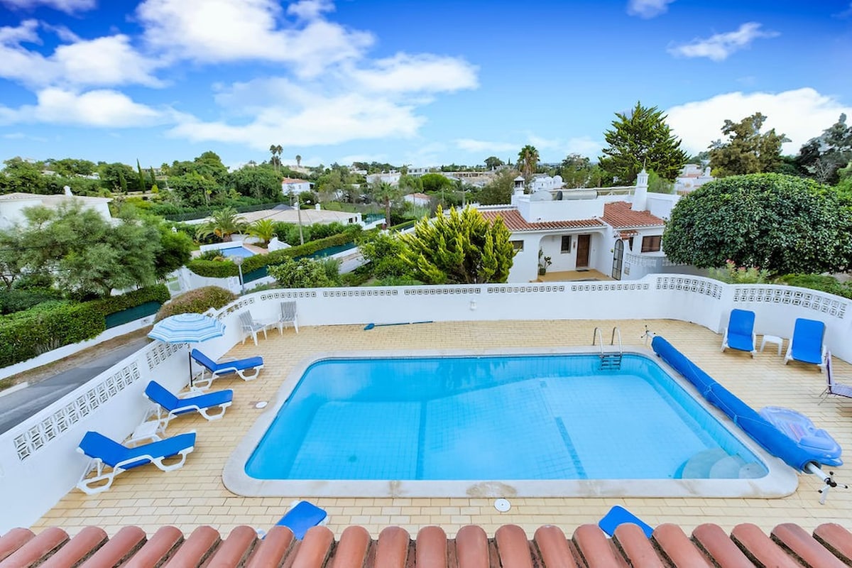 An aerial view of the spacious pool area showcases a large swimming pool surrounded by sun loungers and a patio. Lush greenery and residential structures can be seen in the background under a clear blue sky.