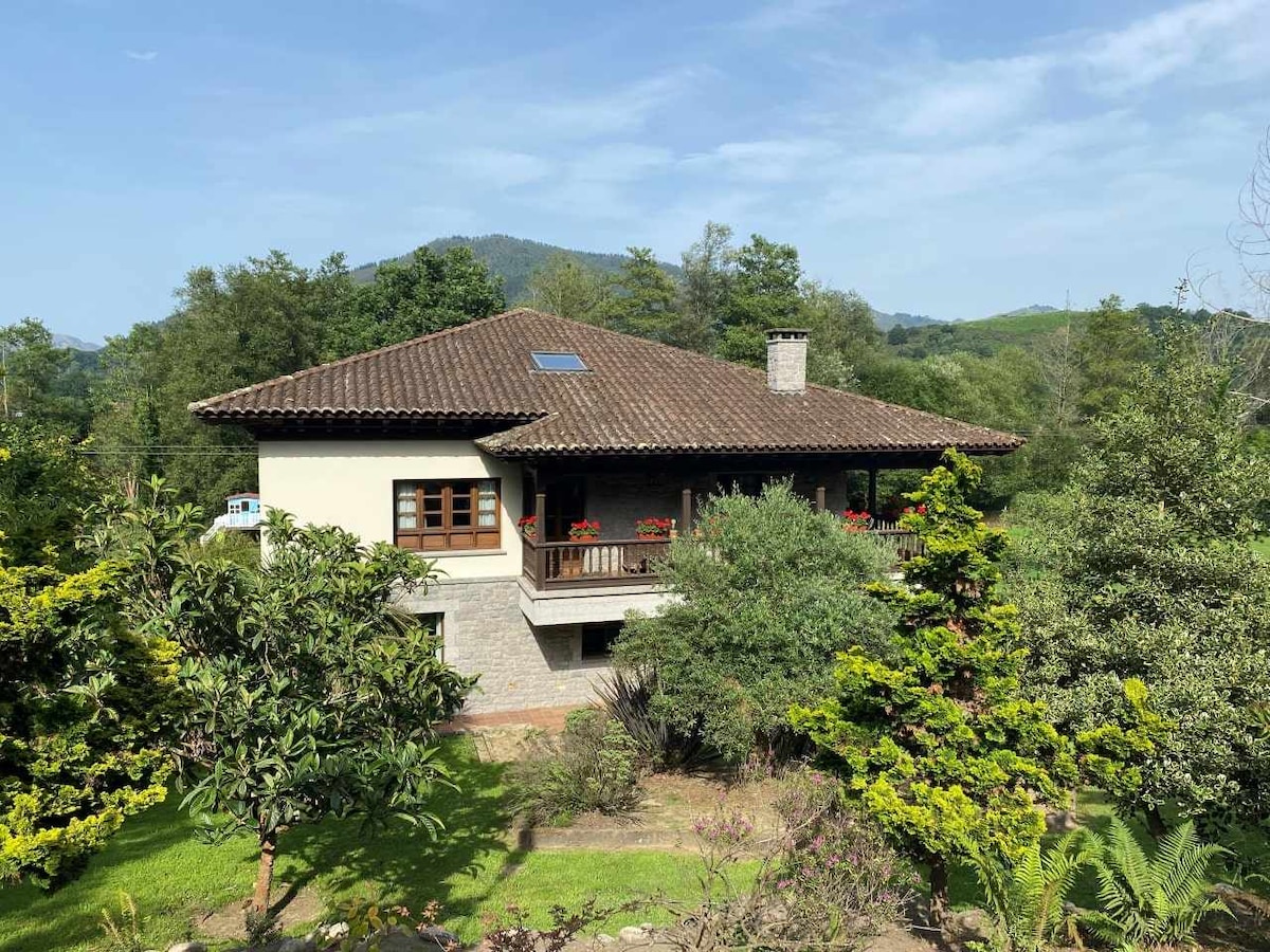 A two-story rural house is surrounded by lush greenery, featuring a brown tiled roof and an inviting front porch with floral arrangements. The scenic backdrop includes distant hills, contributing to the tranquil atmosphere of the estate.
