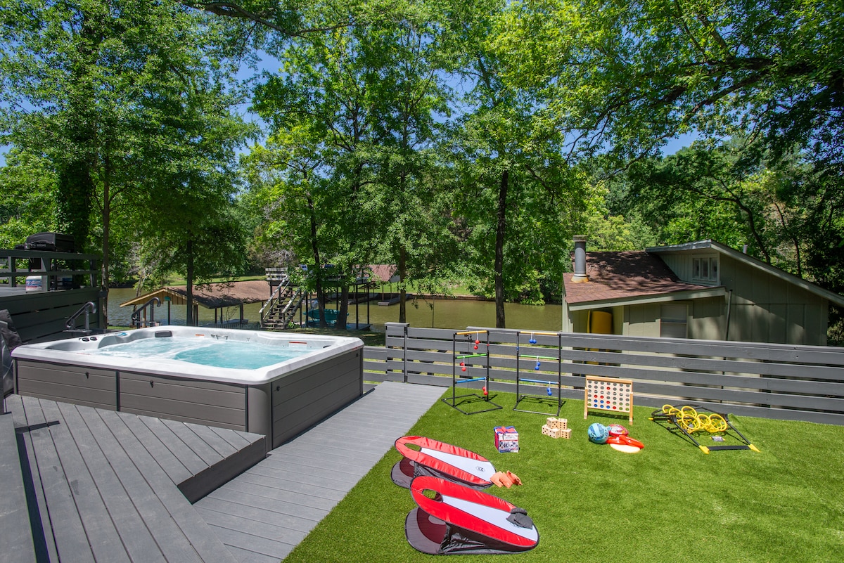 An outdoor deck features a swim hot tub surrounded by lush greenery. Colorful lawn games, including a set of cornhole and ring toss, are set on the artificial grass. In the background, a serene view of Cedar Creek Lake and a separate guest house are visible.