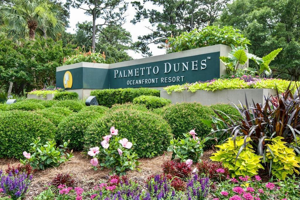 The entrance to Palmetto Dunes Oceanfront Resort is featured, showcasing neatly trimmed shrubbery and vibrant flower beds. The resort sign is prominently displayed against a backdrop of lush greenery, inviting guests to explore the area.