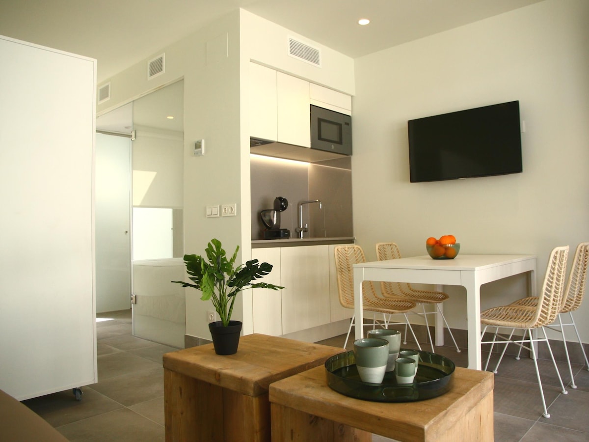 A modern open kitchen area is complemented by a dining table surrounded by four wicker chairs. A small plant is positioned on a wooden coffee table, and a wall-mounted television is visible above the kitchen counter. Natural light brightens the space through a nearby glass door.
