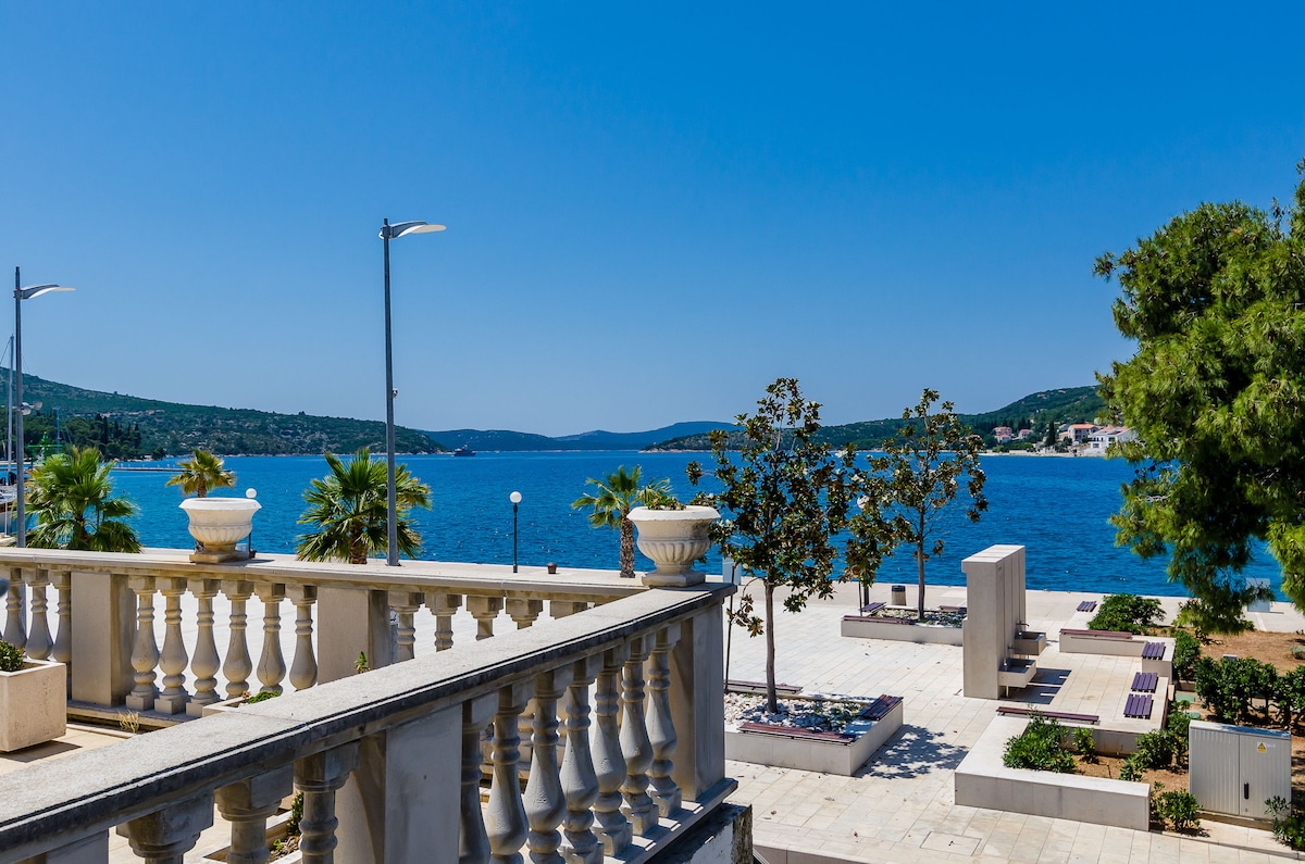 The image showcases a serene waterfront view, featuring clear blue waters and lush greenery along the promenade. A spacious paved area is visible, lined with decorative planters and palm trees, creating an inviting outdoor space. The backdrop includes distant hills under a clear blue sky.