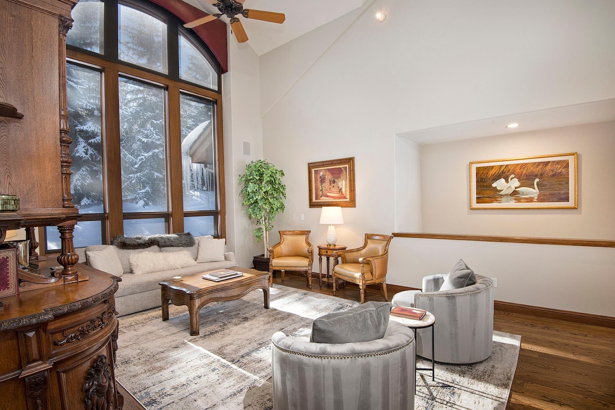 A well-lit living area features large windows showcasing snow-covered trees outside. Comfortable seating includes a plush sofa and two inviting armchairs around a wooden coffee table. A piece of artwork adorns the wall, complemented by a decorative plant in one corner.