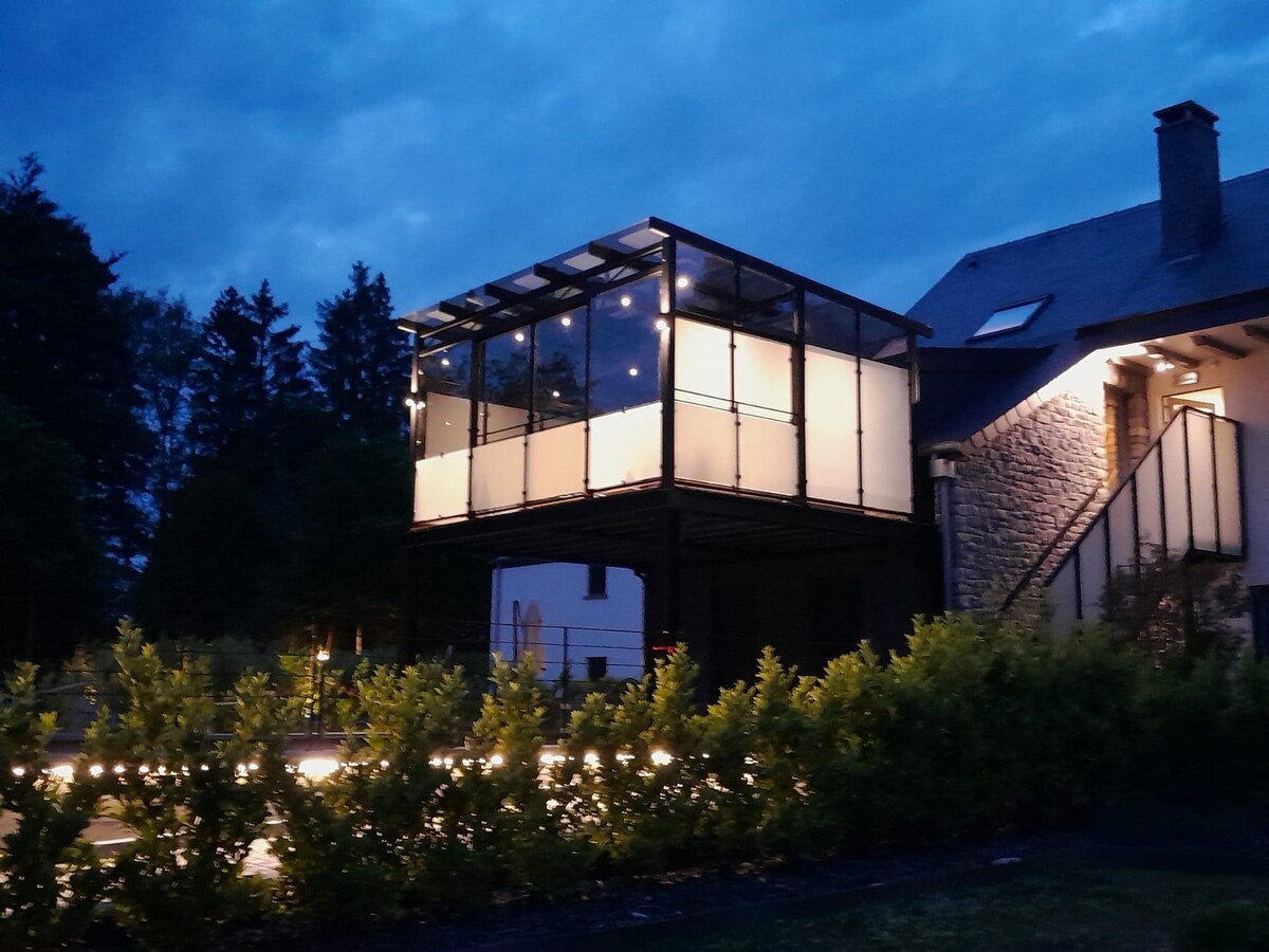 A modern structure is illuminated at dusk, showcasing a glass-enclosed terrace with a patio. Soft lighting enhances the space, surrounded by lush greenery. The house features an exterior of stone and wood, complemented by trees in the background, creating a serene environment.