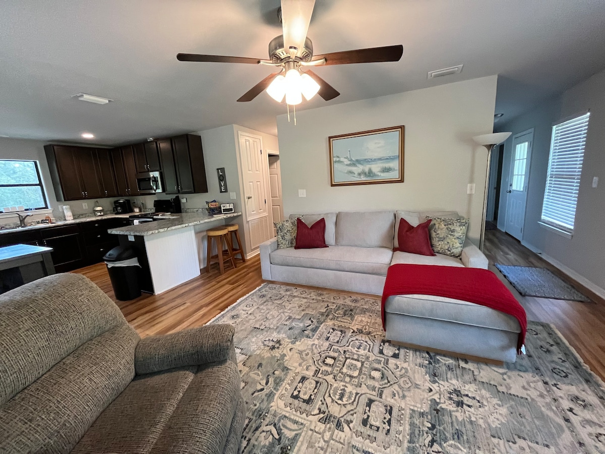 A comfortable living area features a light gray sectional sofa adorned with red and patterned cushions. A cozy armchair is positioned nearby, alongside a large area rug. Light wood flooring is complemented by a ceiling fan, with a view of the kitchen area visible in the background.