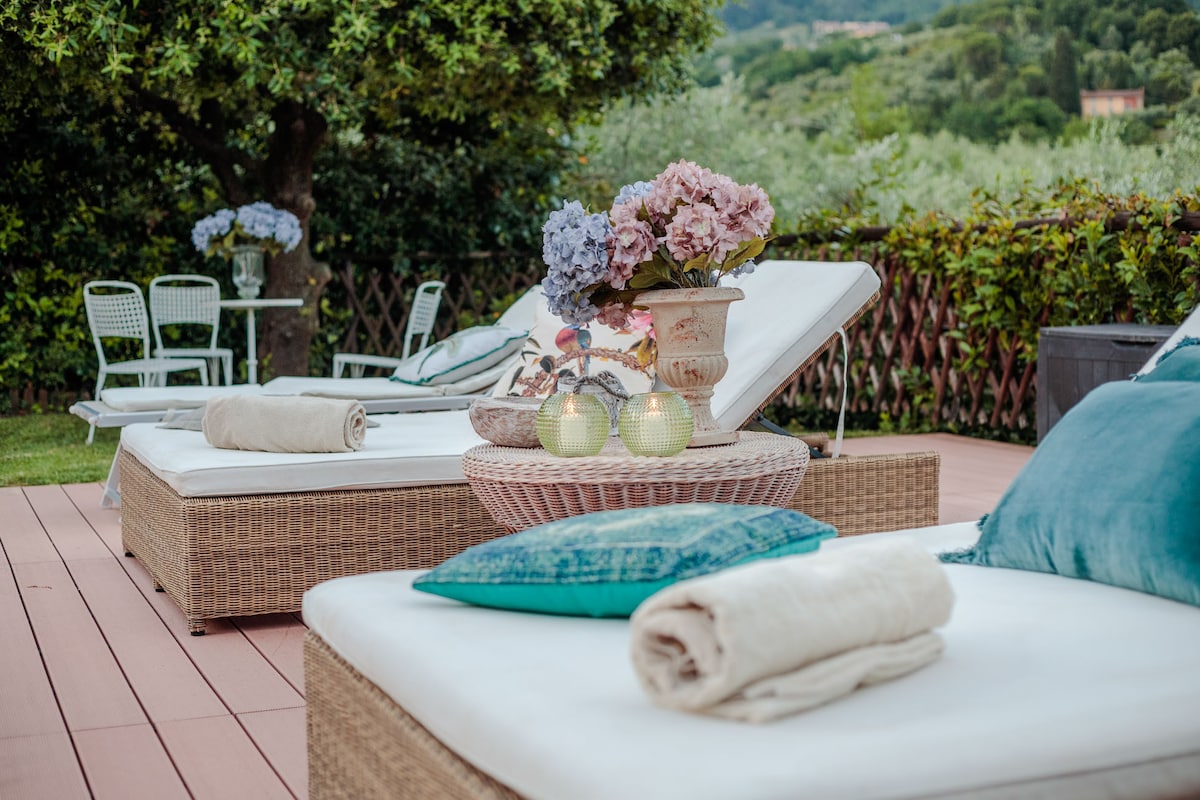 Two sun loungers with plush cushions are arranged on a wooden deck, accompanied by rolled towels. A decorative table features a floral arrangement of hydrangeas. In the background, a small seating area is visible, surrounded by lush greenery and olive trees.