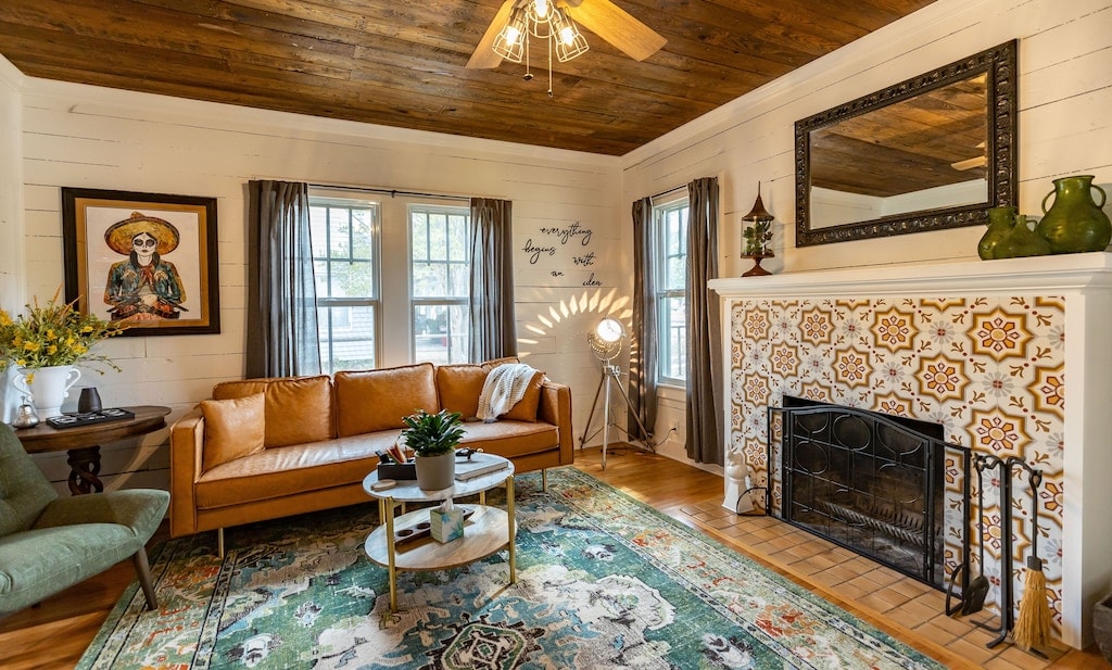 The living area features a warm color palette with a brown leather sofa, green accent chair, and a patterned area rug. Natural light fills the space through large windows, while a decorative fireplace and unique tile work add character. A ceiling fan is present, enhancing comfort.
