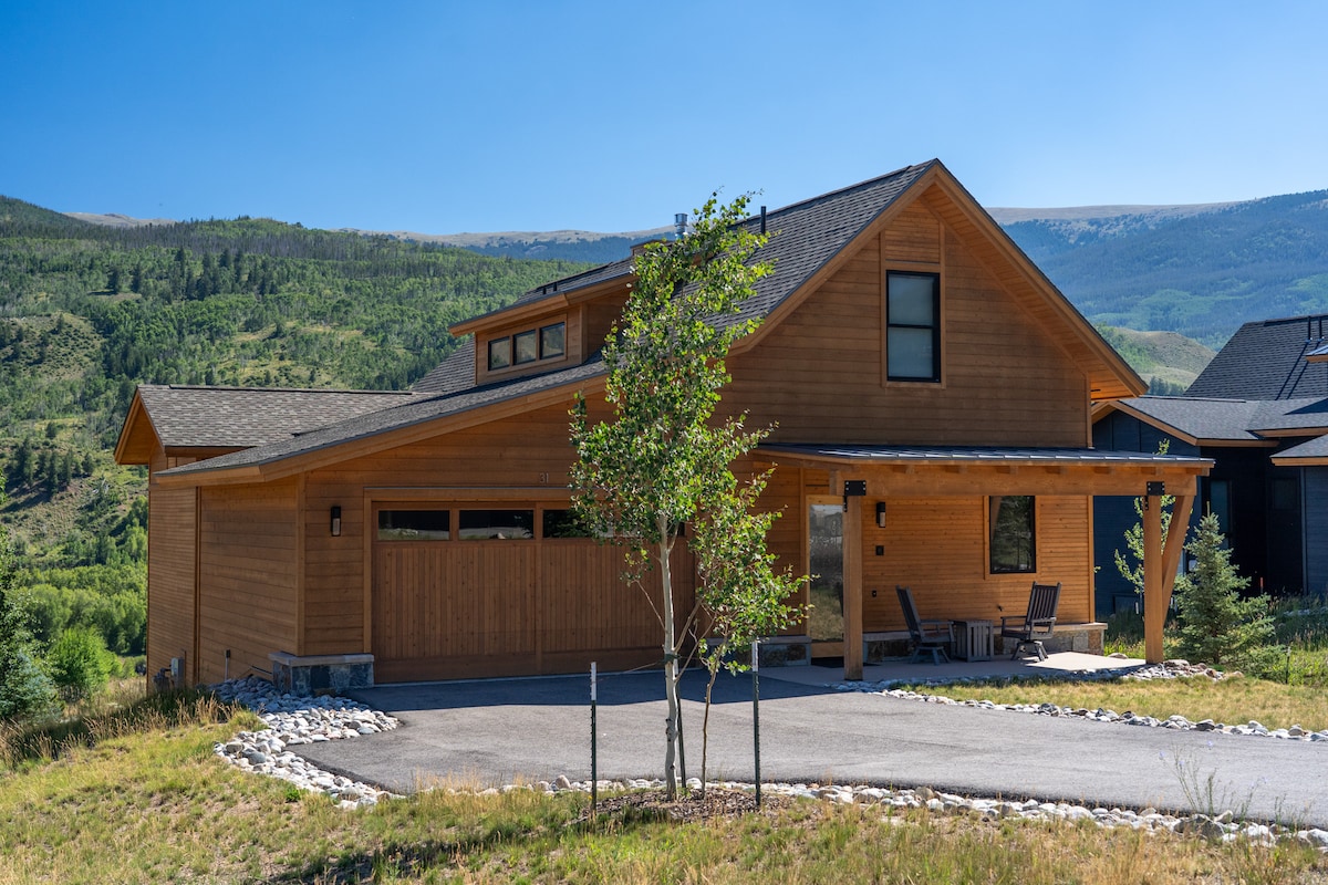 A modern wooden home is situated in a lush green setting, featuring a spacious driveway and a covered porch with seating. The exterior showcases a blend of natural materials, complemented by the surrounding mountains under a clear blue sky.