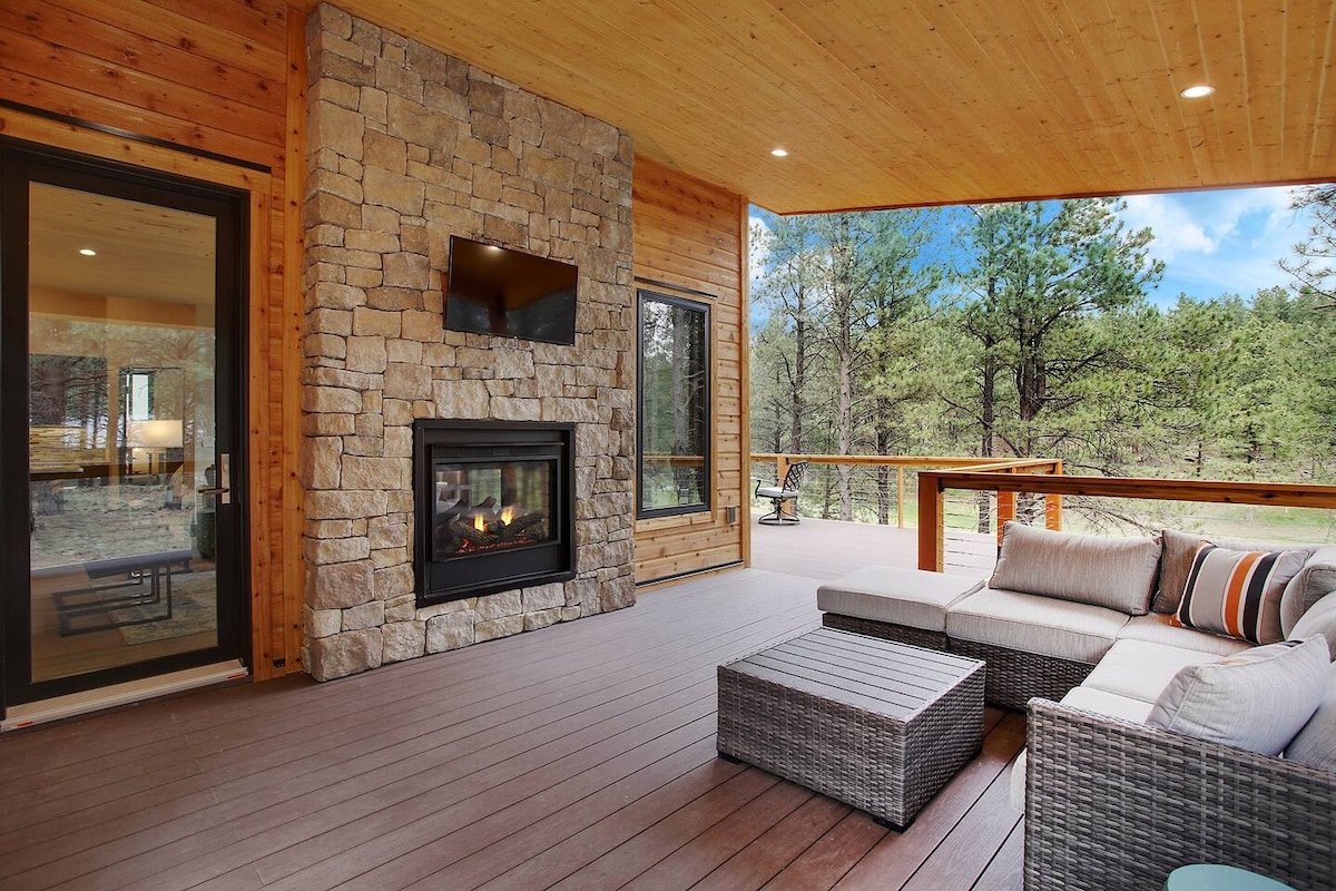 An inviting outdoor seating area features a modern sectional with soft cushions. A stone fireplace is centrally located on the wall, while a wooden bar table stands nearby. Framed by large windows, the space offers views of surrounding greenery.