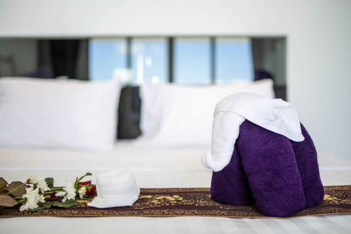 A towel elephant is arranged on a decorative runner, accompanied by fresh flowers. Soft white linens cover the spacious king-size bed, with large windows in the background allowing natural light to fill the room.