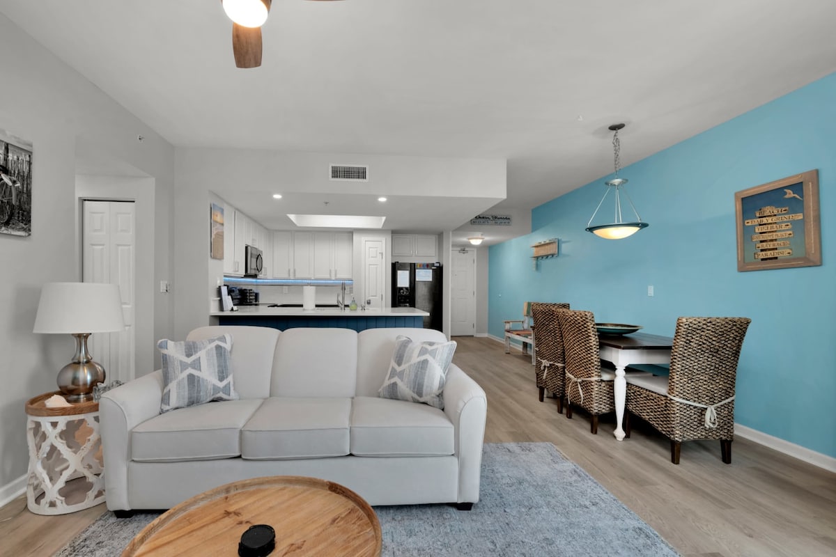 A bright and open living space is observed, featuring a comfortable sofa with decorative cushions. A wooden coffee table sits in front of the sofa. Beyond, a dining area is highlighted with a white table surrounded by woven chairs, set against a light blue accent wall.