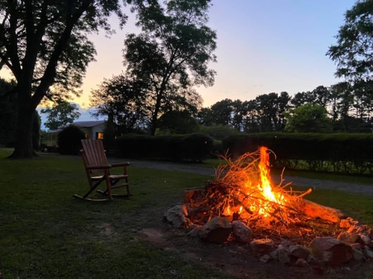 A fire pit created with stones is surrounded by a grassy area, featuring a burning fire with glowing orange and yellow flames. A wooden rocking chair is positioned nearby, framed by trees and the lush landscape at dusk.