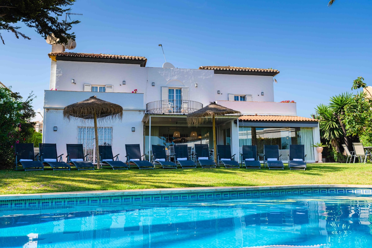 A spacious villa is showcased with multiple lounge chairs arranged along a lush green lawn, all facing an inviting pool. The exterior features a clean facade, balconies, and a shaded seating area under a thatched umbrella. Clear blue skies enhance the leisurely atmosphere.