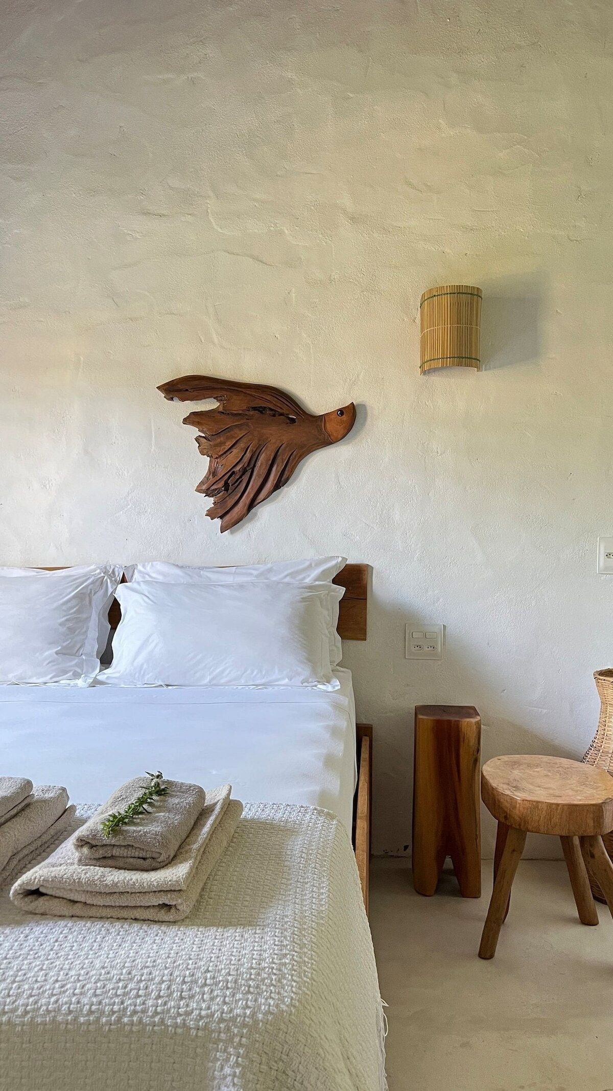 A comfortable sleeping area is highlighted by a rustic wooden bird sculpture on the textured wall. The spacious bed is adorned with crisp white linens, complemented by neatly folded towels on the edge. Adjacent is a natural wood stool and a woven chair, enhancing the serene ambiance.