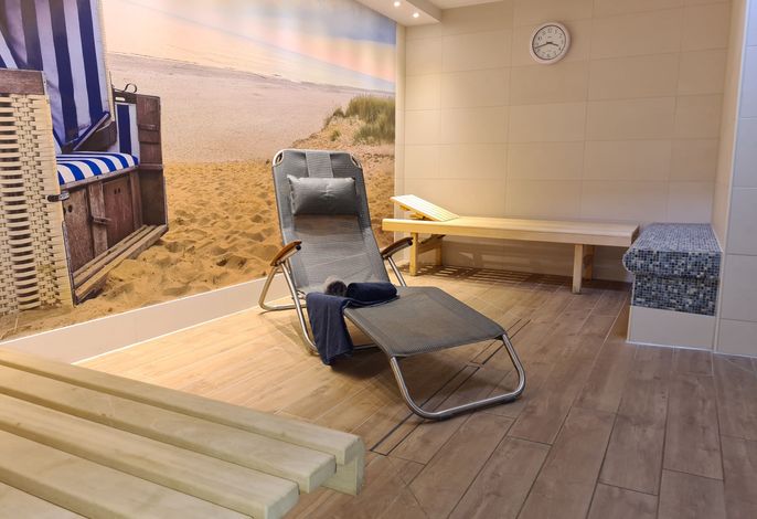 A relaxation area features a comfortable lounger with a cushion, positioned against a sandy beach mural. Wooden benches are arranged along the wall, adding to the calming atmosphere. A clock hangs above, and soft lighting enhances the tranquil setting.