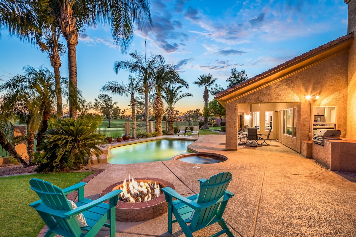 An outdoor space features a heated pool with a water feature and a spa, surrounded by tall palm trees and manicured grass. A built-in barbecue is visible alongside a fire pit with adirondack chairs, all set against a scenic sunset view.