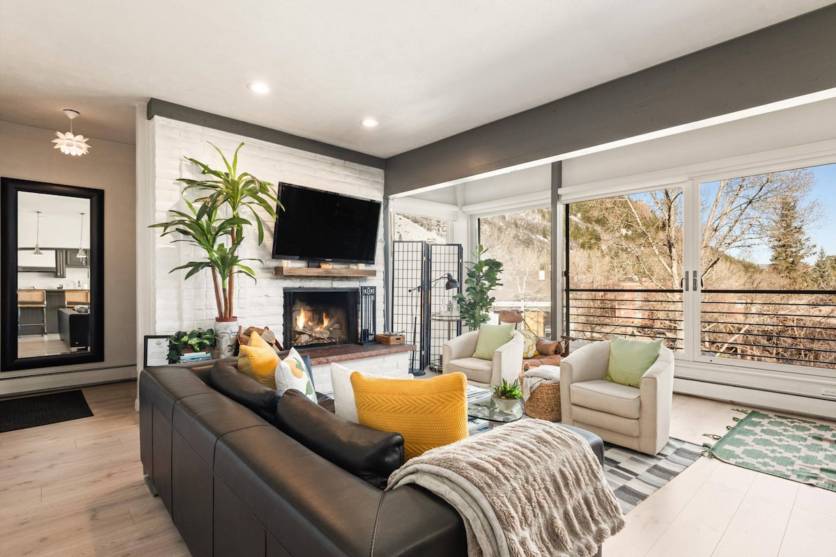 A spacious living area features a modern black leather sectional sofa with colorful accent pillows. A wood-burning fireplace is framed by a white stone wall and complemented by natural light from large floor-to-ceiling windows. Comfortable seating arrangements provide relaxation, with views of the surrounding landscape.