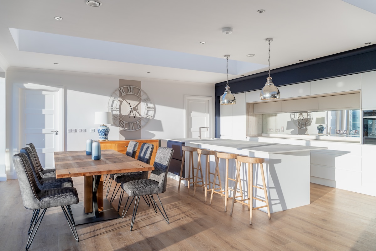A modern open plan kitchen and dining area features a wooden dining table surrounded by six black chairs. A kitchen island with high stools complements glossy cabinetry and stainless steel appliances, while large windows allow natural light to brighten the space.