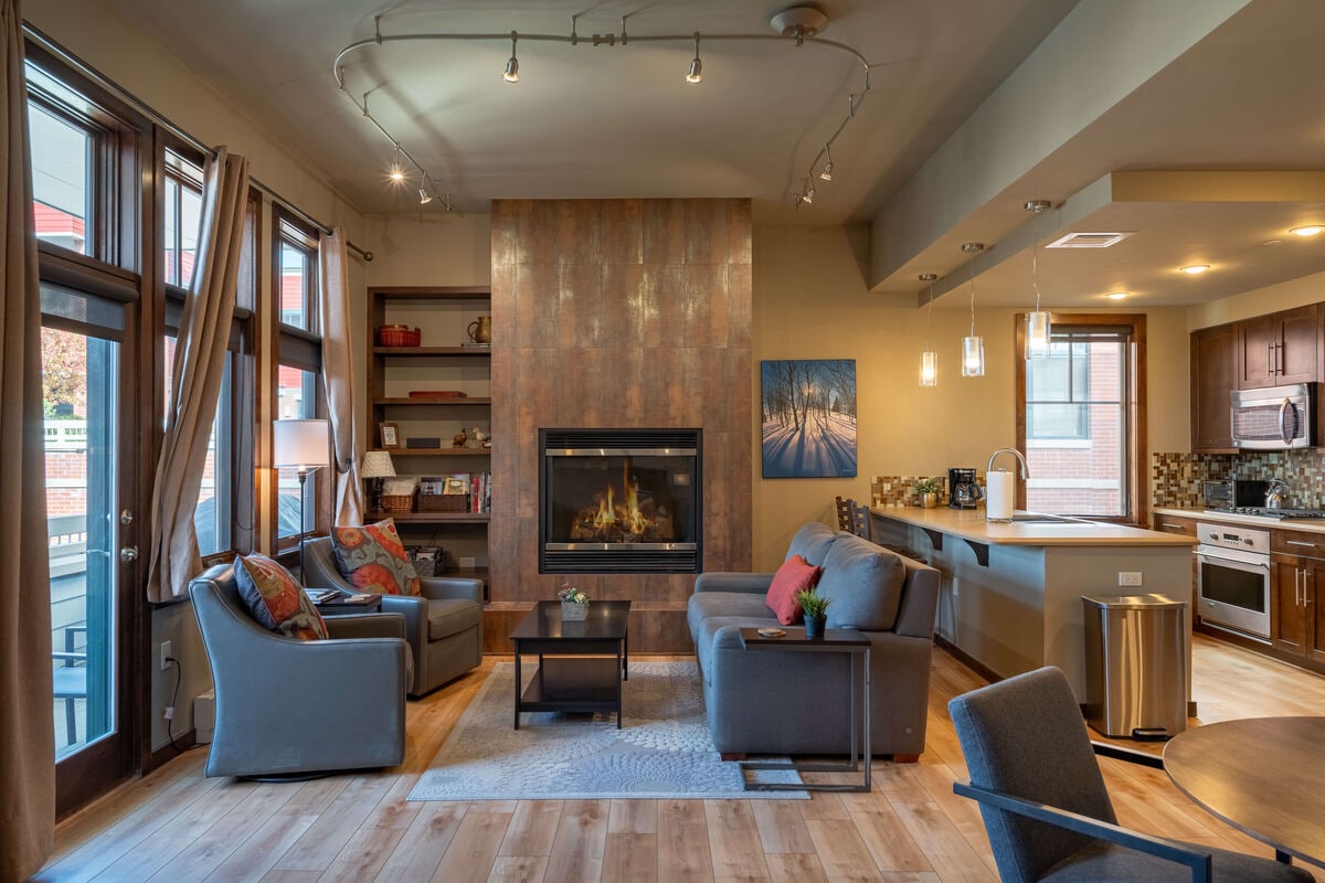 The open living area features a cozy seating arrangement with modern sofas placed around a stylish area rug. A warm fireplace is framed by textured wall panels, while a fully equipped kitchen and dining area are visible in the background, illuminated by pendant lighting.