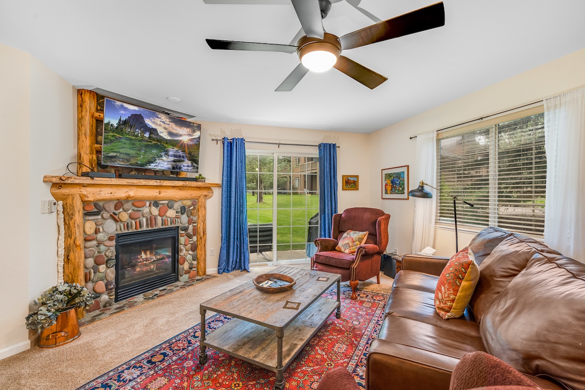 The living area features a large gas fireplace framed by a stone mantle. A smart TV is mounted above, while two armchairs and a spacious sofa provide comfortable seating. Light enters through large windows, with blue curtains adding a pop of color. A rustic coffee table sits on a patterned rug.