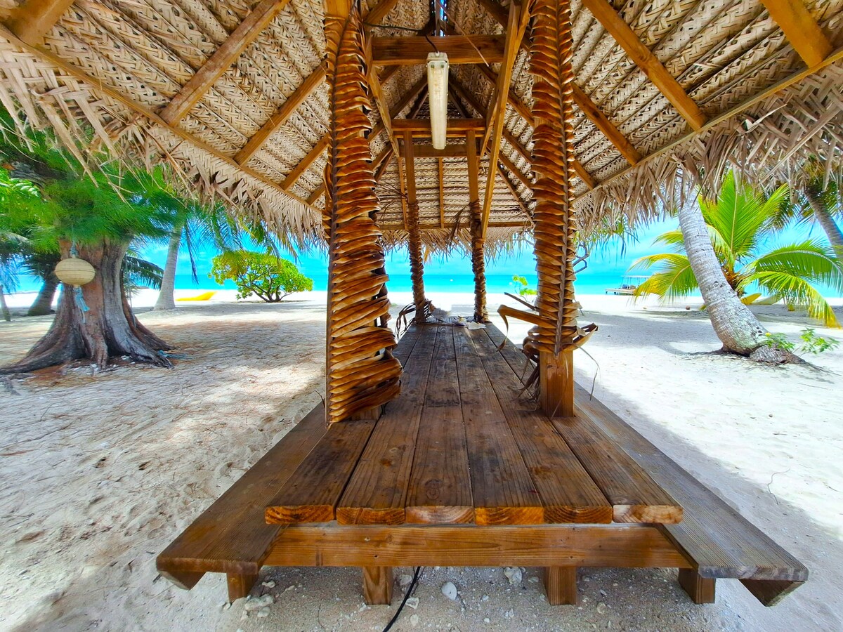 A sheltered wooden seating area stands on a sandy beach, supported by natural palm trunk columns. The thatched roof offers shade while providing views of the turquoise lagoon and lush greenery in the background.