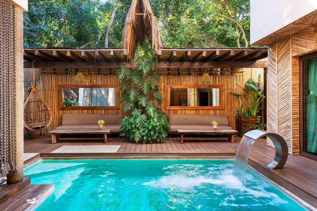 A private outdoor area features a tranquil pool with a stylish water feature. Two wooden lounge beds are framed by tropical greenery, while bamboo details enhance the natural aesthetic. Bright plants and warm wood tones create a serene relaxation space.