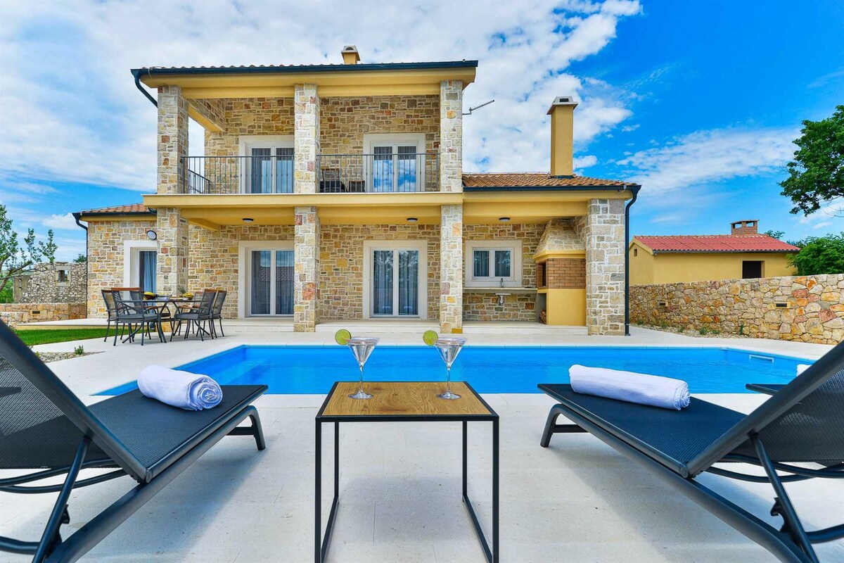 An outdoor area features a sparkling pool surrounded by lounge chairs and a small table, perfect for relaxation. The villa showcases a stone exterior with two stories, large windows, and a balcony overlooking the pool area, set against a partly cloudy sky.
