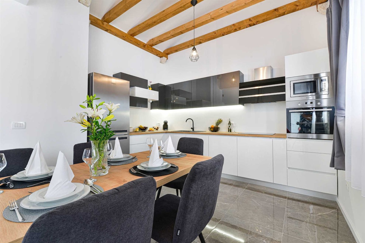 A modern kitchen is highlighted by a spacious dining table set for six, featuring elegant tableware. The sleek cabinetry in dark and light tones complements stainless steel appliances. Natural light floods the space through a large window, enhancing the open and airy atmosphere.