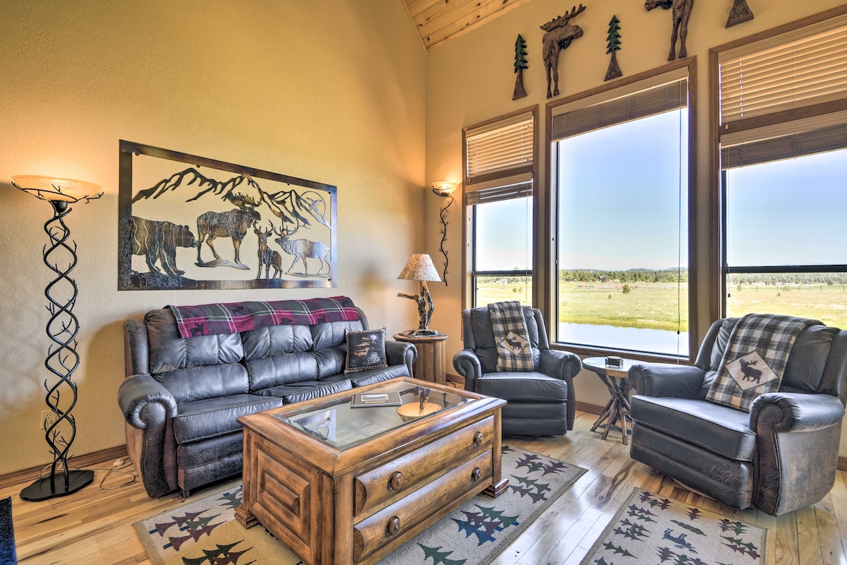 A cozy living room features large windows framing scenic views of the mountains and open fields. A leather couch and two armchairs provide seating, accompanied by a wooden coffee table. Wall decor includes a mountain-themed artwork and wooden animal carvings.