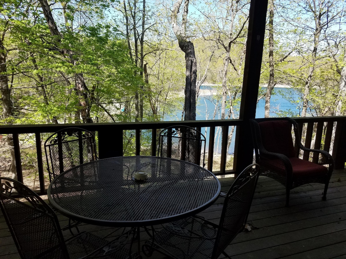 A cozy outdoor deck is furnished with a round metal table surrounded by six chairs. A red cushioned armchair is positioned nearby, providing a comfortable seating option. Lush green trees frame the view of the tranquil lake, visible through the open railing.
