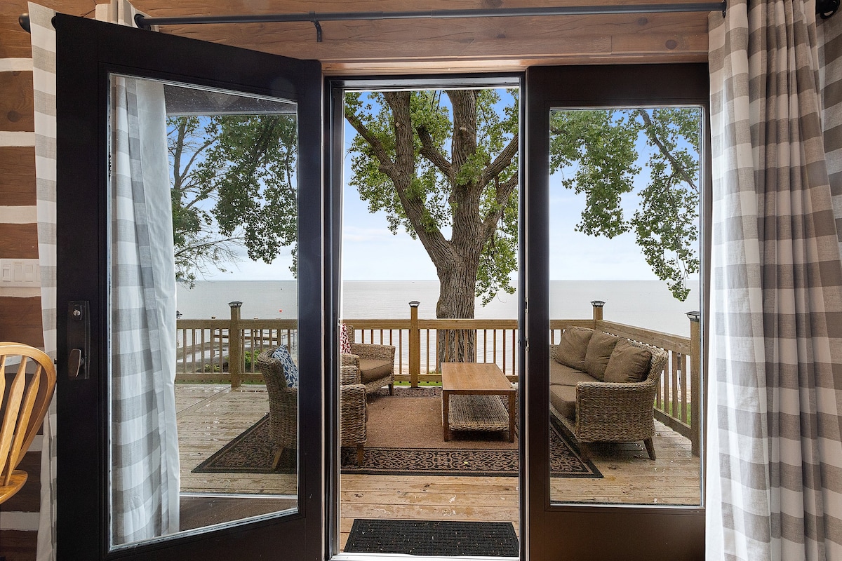 Glass doors frame a view of the waterfront, showcasing a spacious outdoor deck with comfortable seating. A large tree provides shade, and the horizon features the tranquil waters of the bay, creating a serene setting for relaxation.