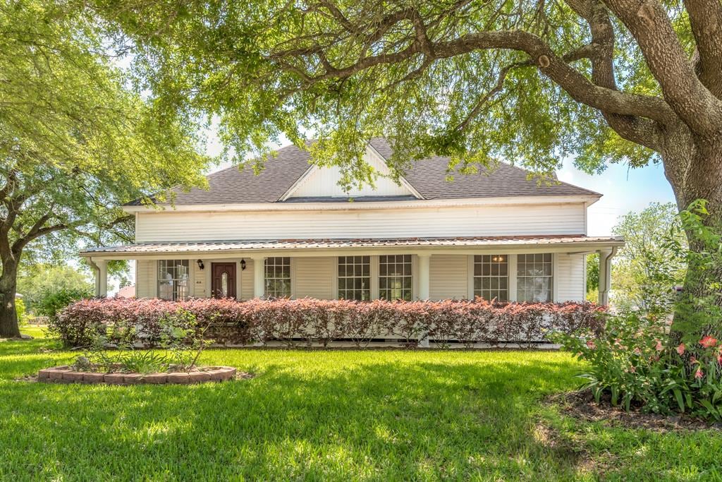 The charming two-story home presents a welcoming front facade, featuring a large porch bordered by manicured shrubs and vibrant landscaping. The exterior is accented by wide windows that allow natural light, with shaded areas provided by mature trees in the yard.