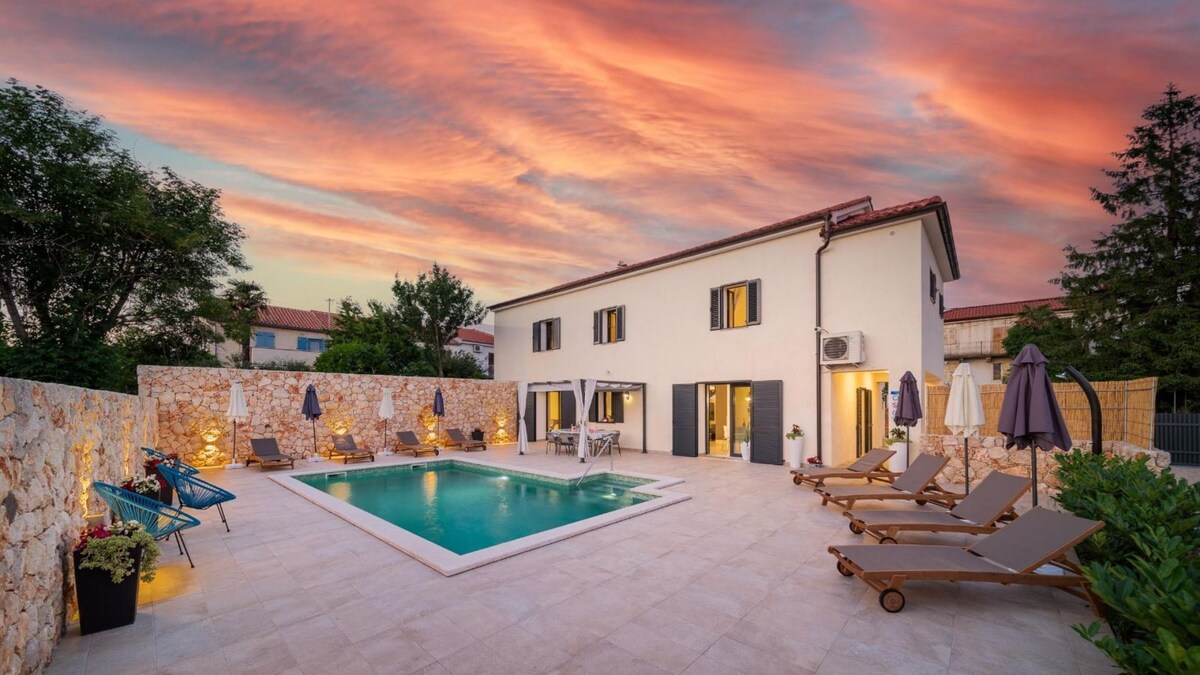An outdoor area features a private pool surrounded by loungers and umbrellas, with vibrant colors lighting up the sky at sunset. The villa stands nearby, showcasing large windows that open to the pool area and a stone wall providing privacy.