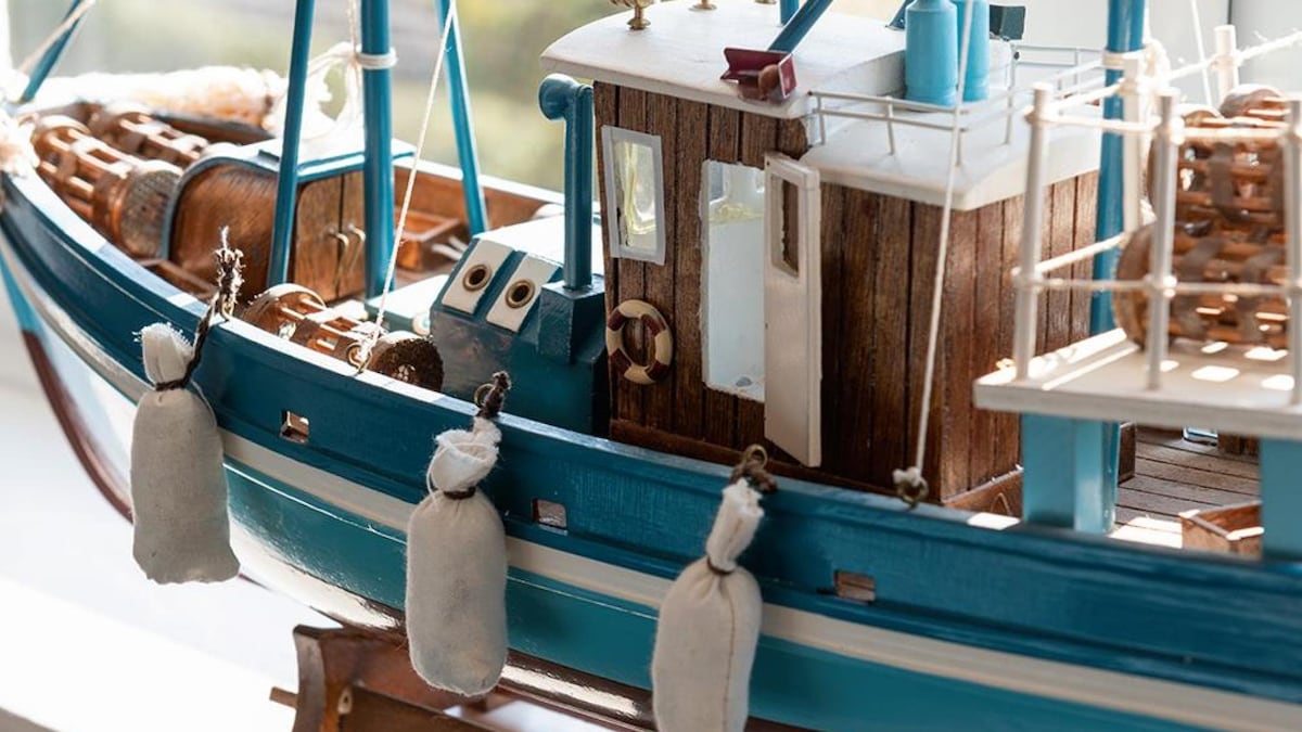 A detailed model of a fishing boat is showcased, featuring a blue hull and wooden accents. The structure has a small cabin with windows, and fishing gear is visibly arranged on deck. Weighted sacks hang from the side, enhancing the realistic appearance of the vessel.